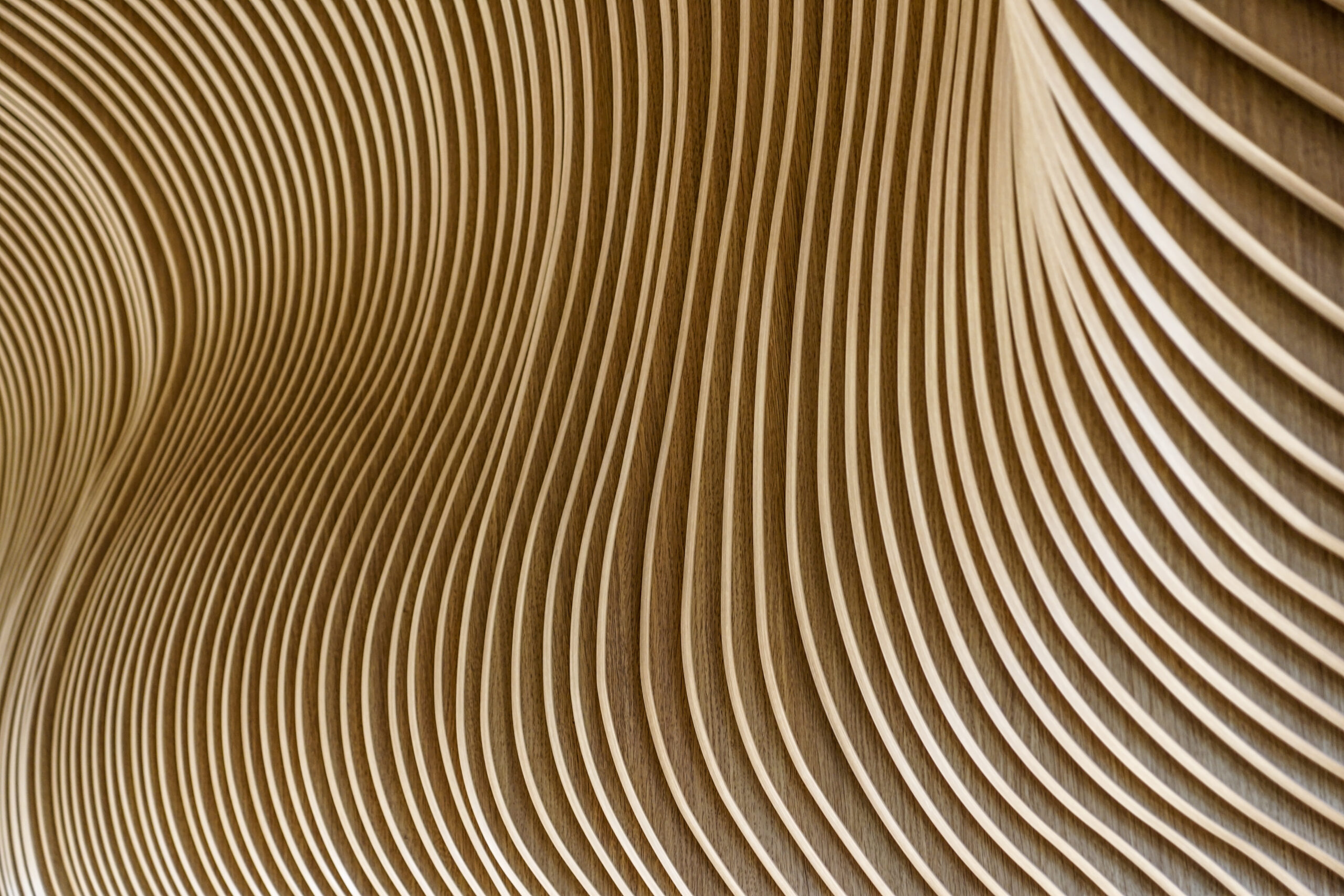 Curved, parallel wooden slats create a smooth, undulating pattern on a wall, forming an abstract, wavy texture with warm, natural tones.