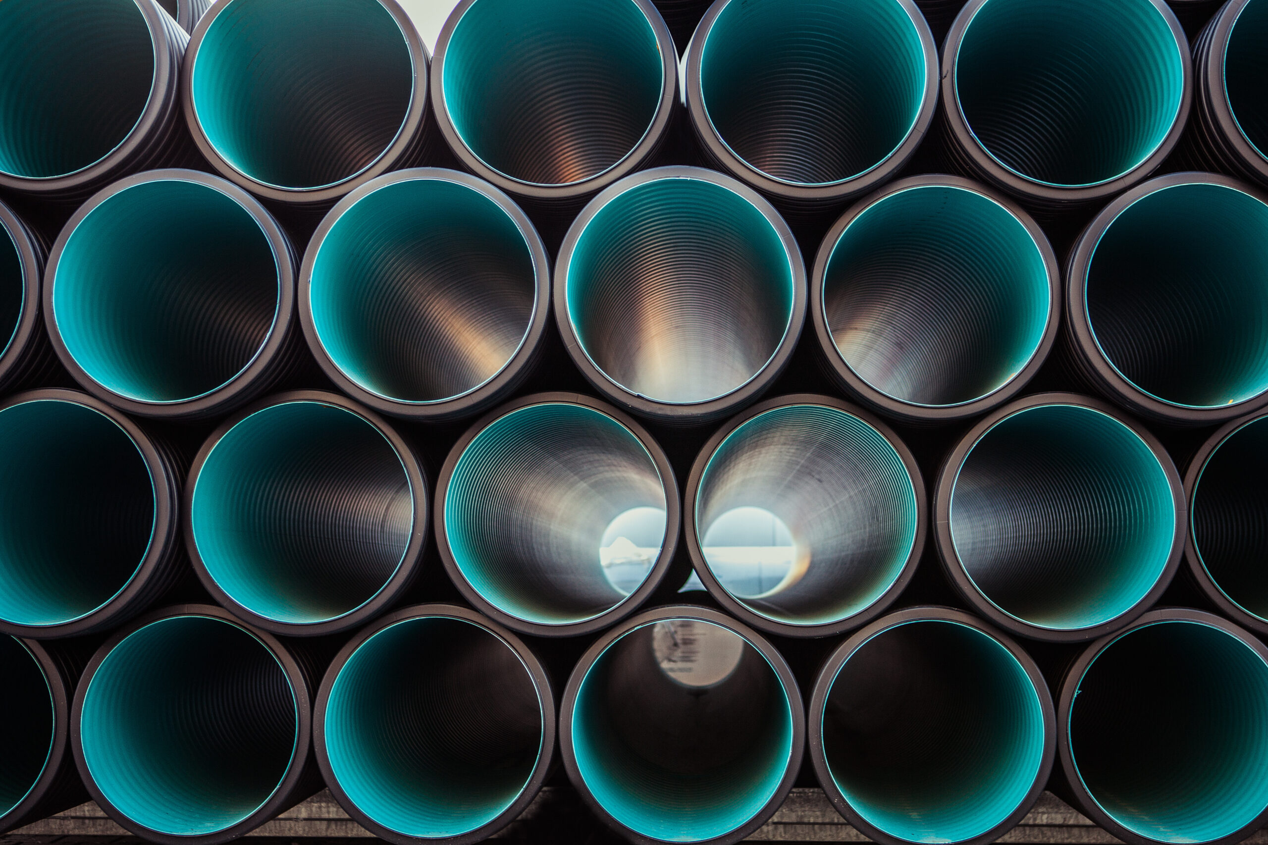 Close-up view of stacked large industrial pipes, arranged in rows. The interiors of the pipes are teal, and light is visible shining through the openings, creating a tunnel-like effect.